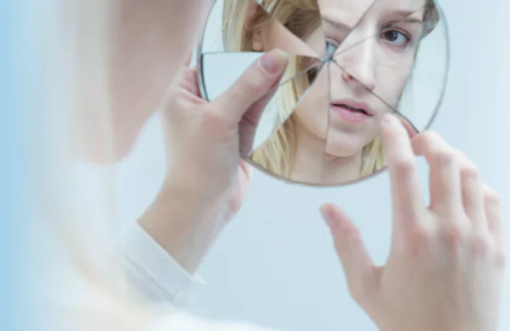 Omslagafbeelding Spiegeling van zelfbeeld. Blonde jonge vrouw die in gebroken spiegel kijkt.